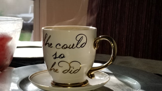 White china oversize coffee cup, fillled with Ma' Cline's delicious coffee, on a saucer with gold handle and gold text "She could so she did" Cup has coffee vapor rising above