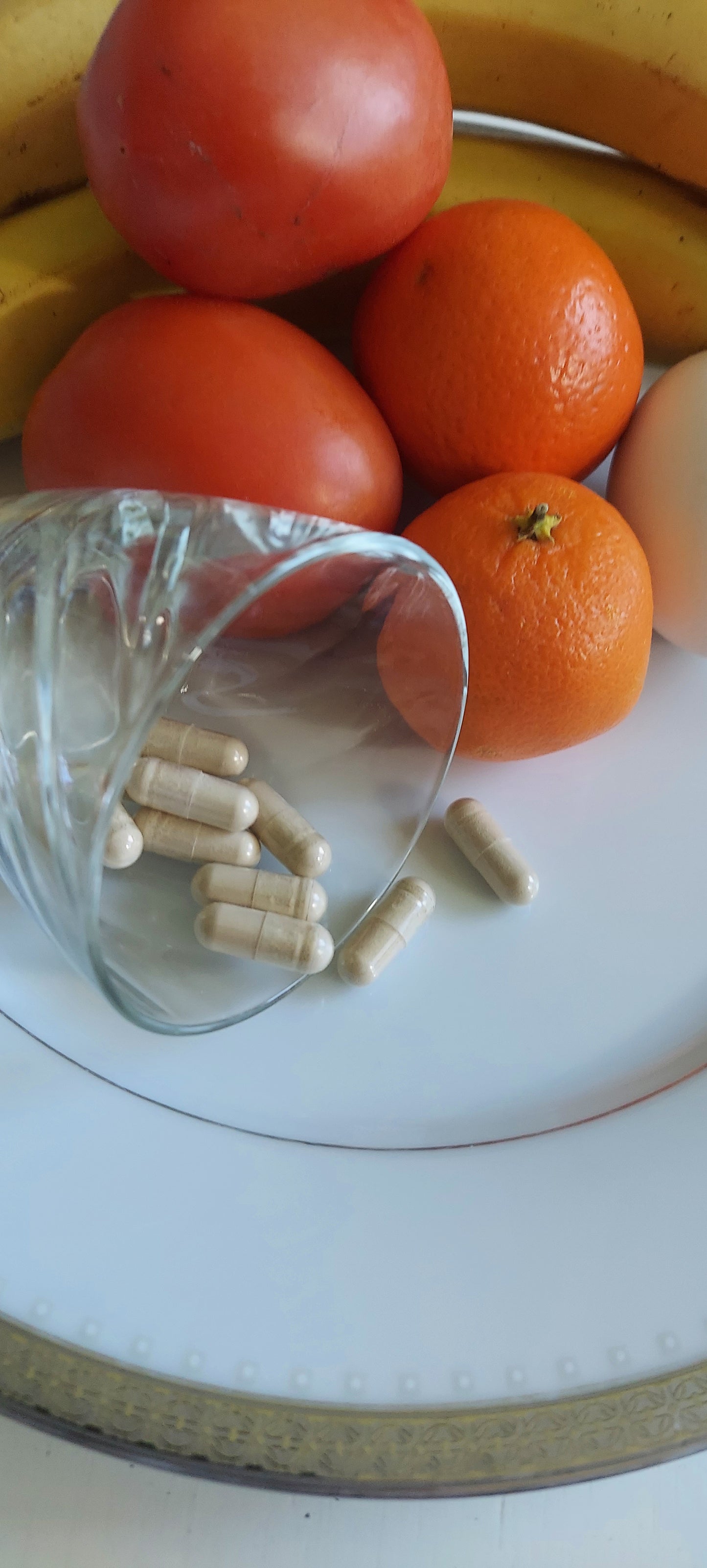 Plate of ripe tomatoes, orange, bananas, and a lemon, also containing a small juice glass on its side spilling food capsules onto the plate with the fruit.