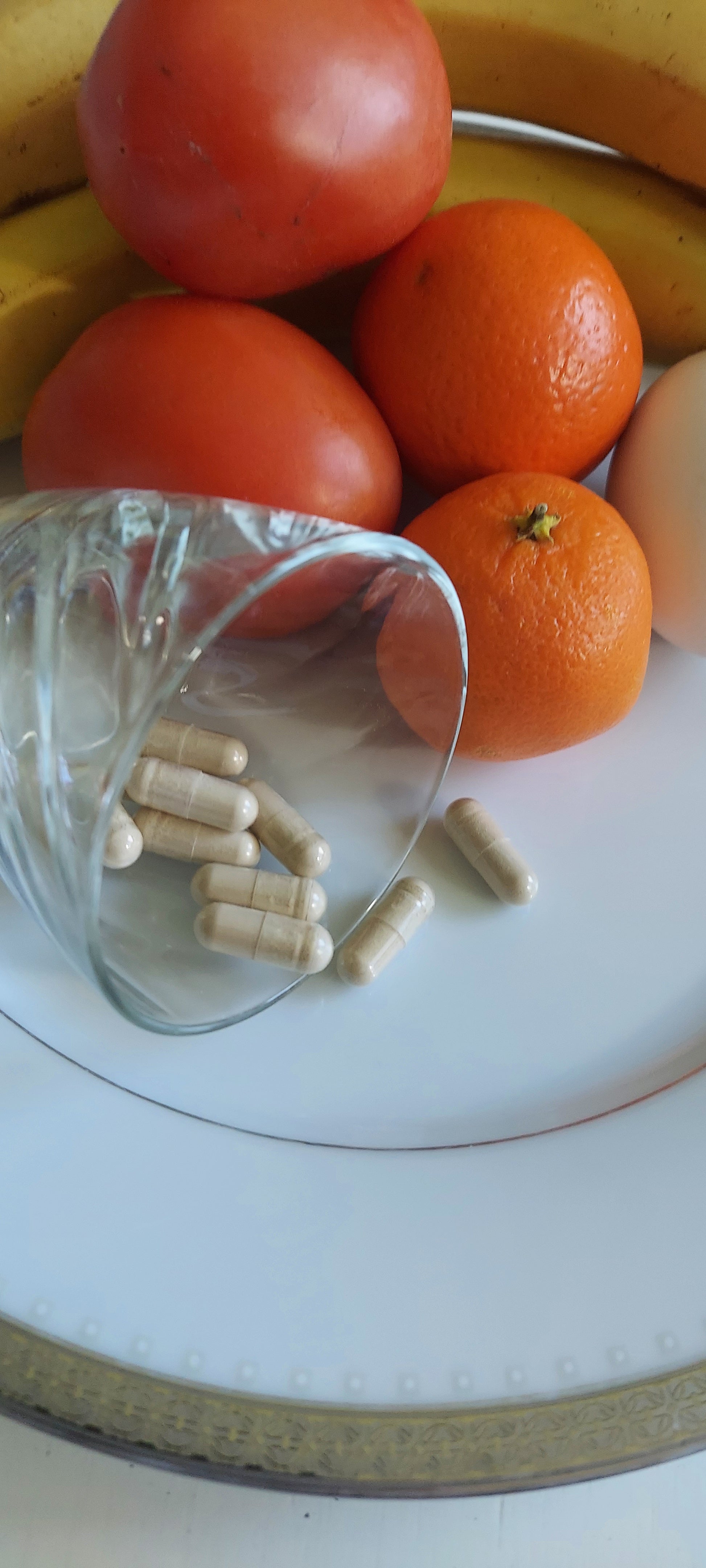 Plate of ripe tomatoes, orange, bananas, and a lemon, also containing a small juice glass on its side spilling food capsules onto the plate with the fruit.