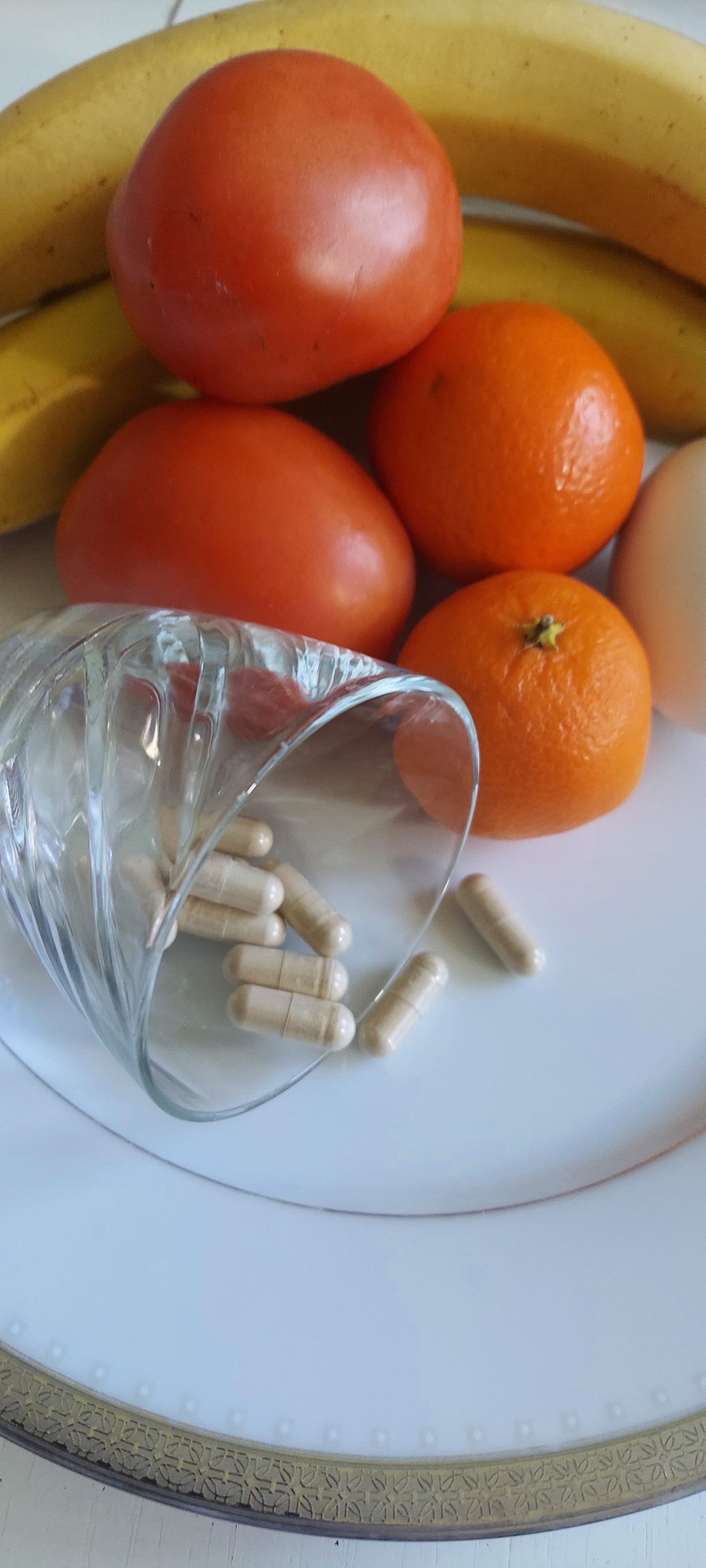 Plate of ripe tomatoes, orange, bananas, and a lemon, also containing a small juice glass on its side spilling food capsules onto the plate with the fruit.