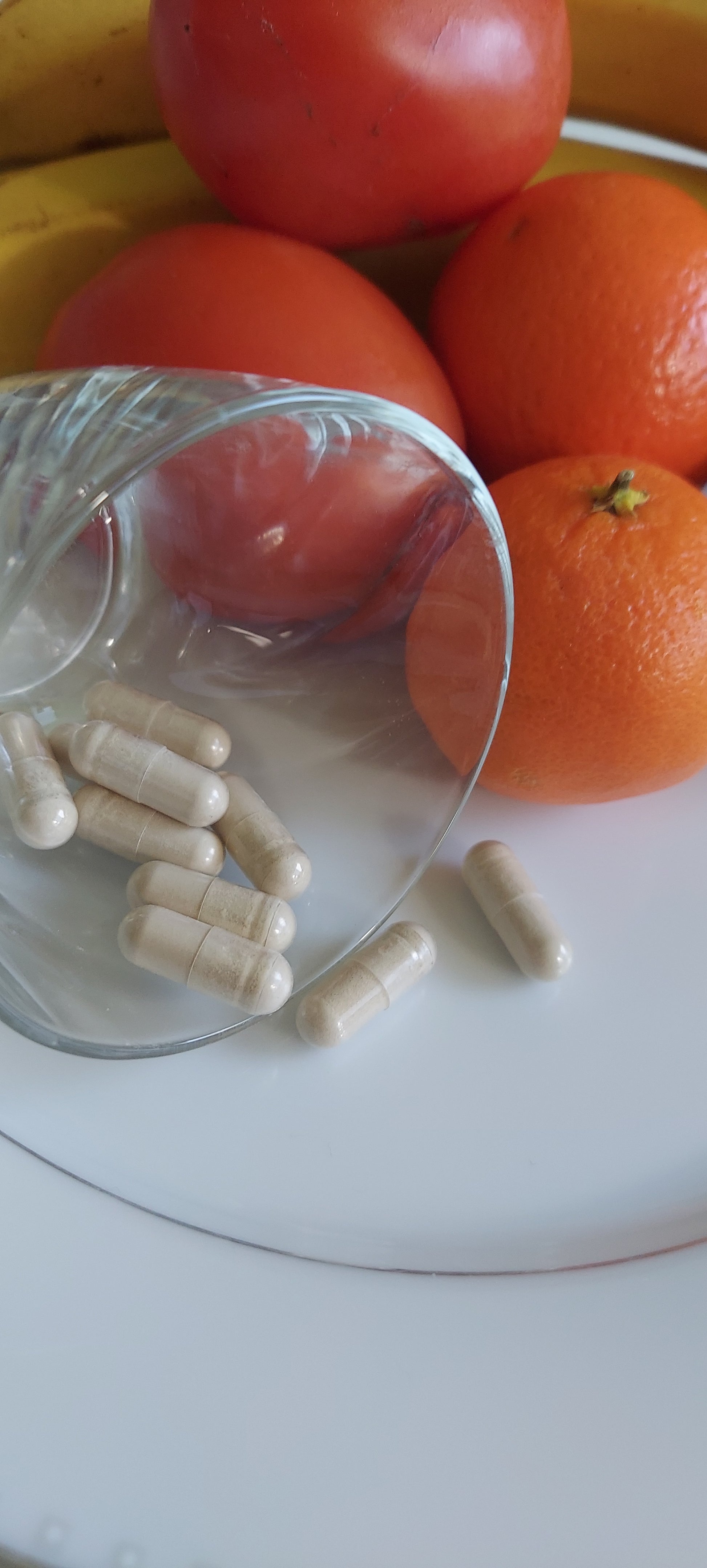 Plate of ripe tomatoes, orange, bananas, and a lemon, also containing a small juice glass on its side spilling food capsules onto the plate with the fruit.