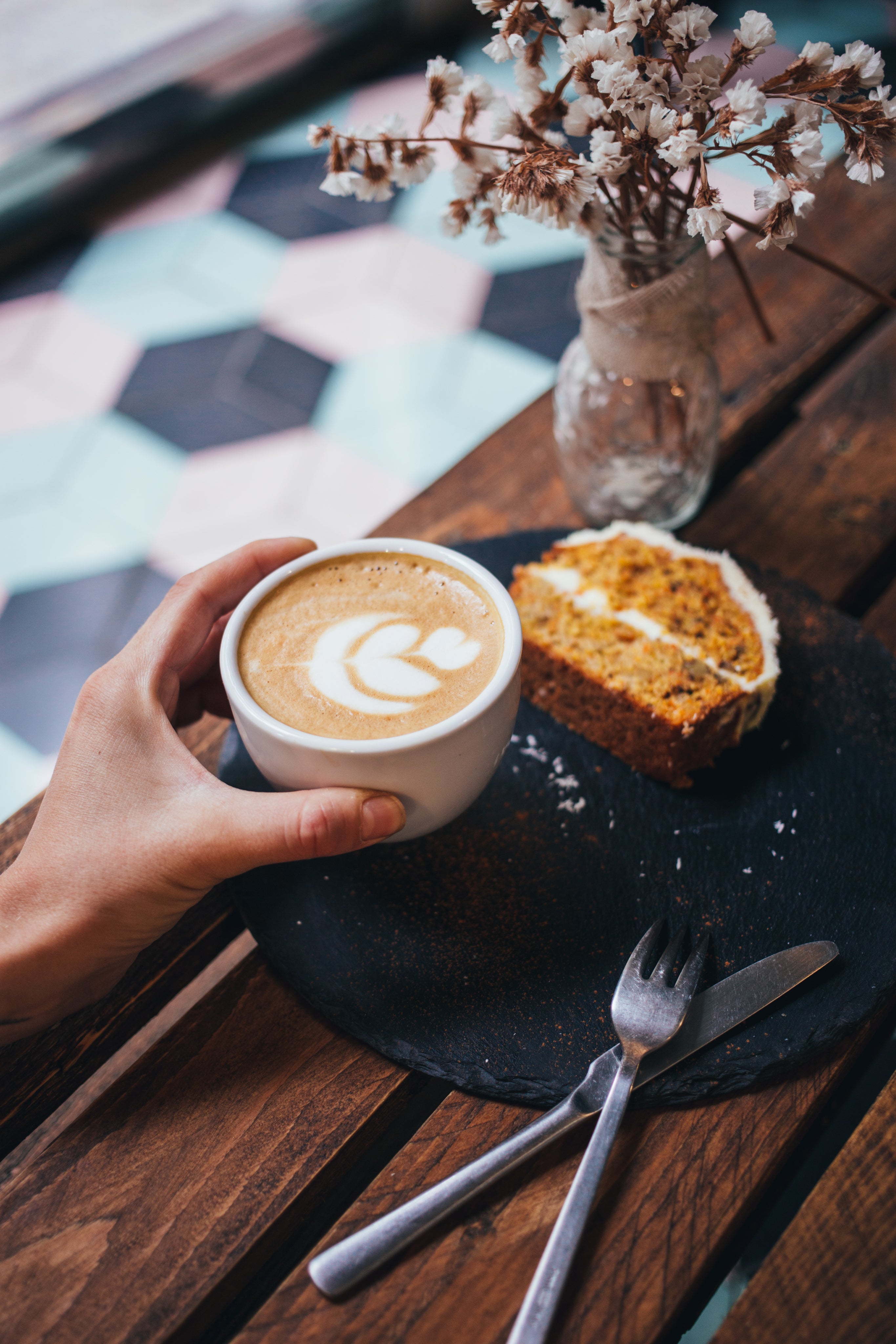 Lady's hand holding a white cup of delicious Ma' Cline's Coffee with cream topper in the shape of a tree. Next to the cup is a slice of iced banana nut bread.