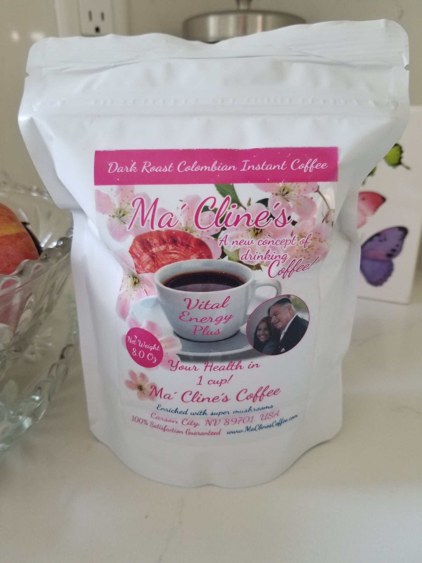 White bag with pink toned front label of Ma' Cline's Vital Instant EnergyPlus Coffee. Background photo is pink cherry blossoms, and red ganoderma mushrooms behind a white cup of delicious, steaming coffee.