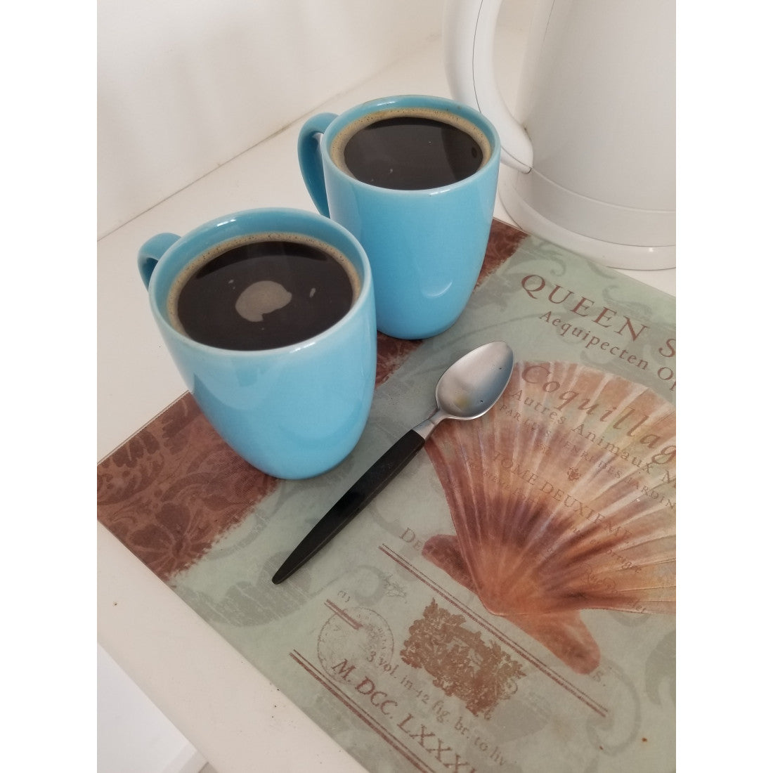 Two sky-blue coffee mugs with simmering Ma' Cline's Coffee on a placemat with a spoon.