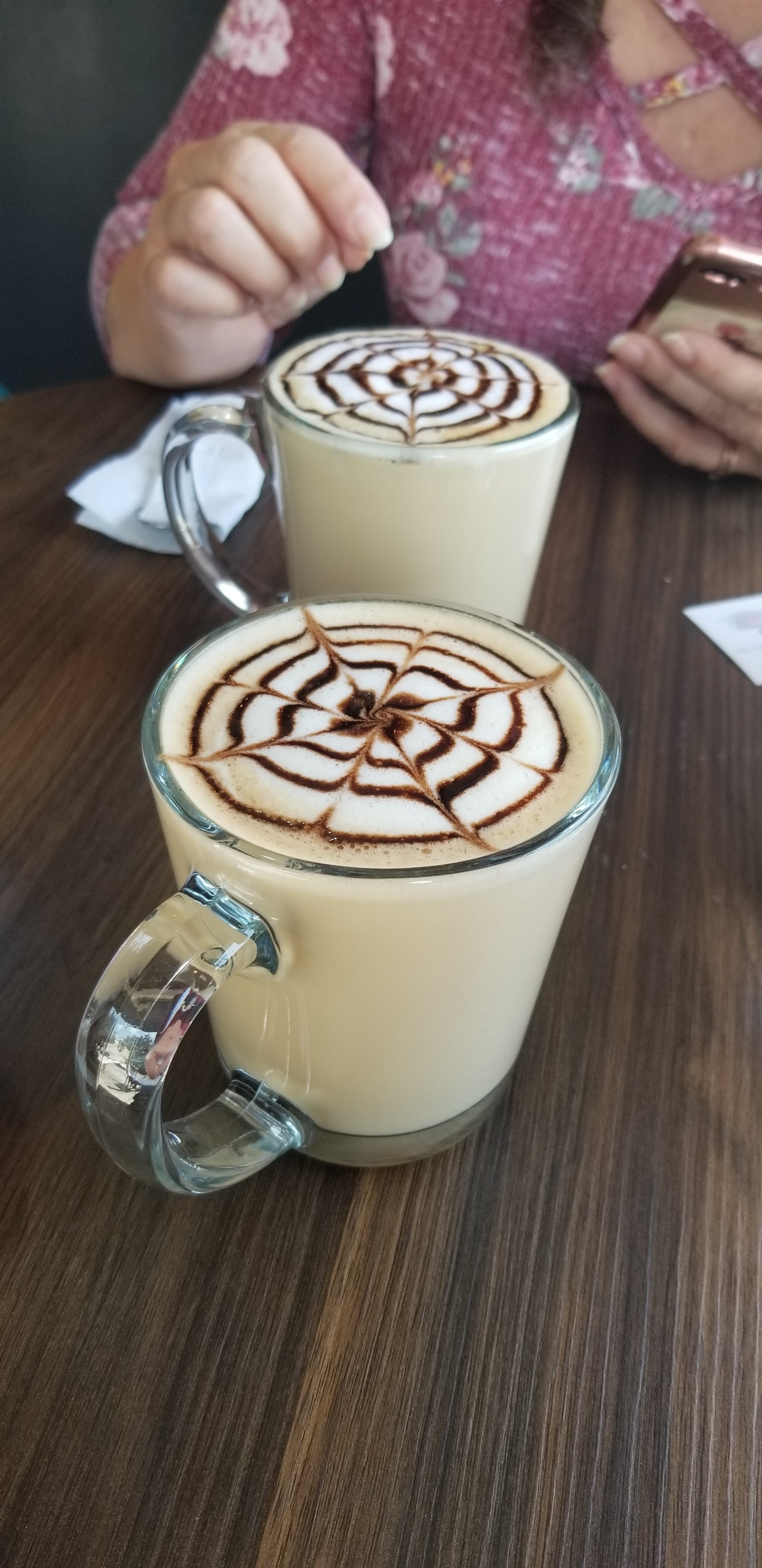 Two transparent glass coffee cups with light tan Ma' Cline's ChocoShroom  Hot Chocolate, wit a decorative chocolate syrup design floating on the top. 