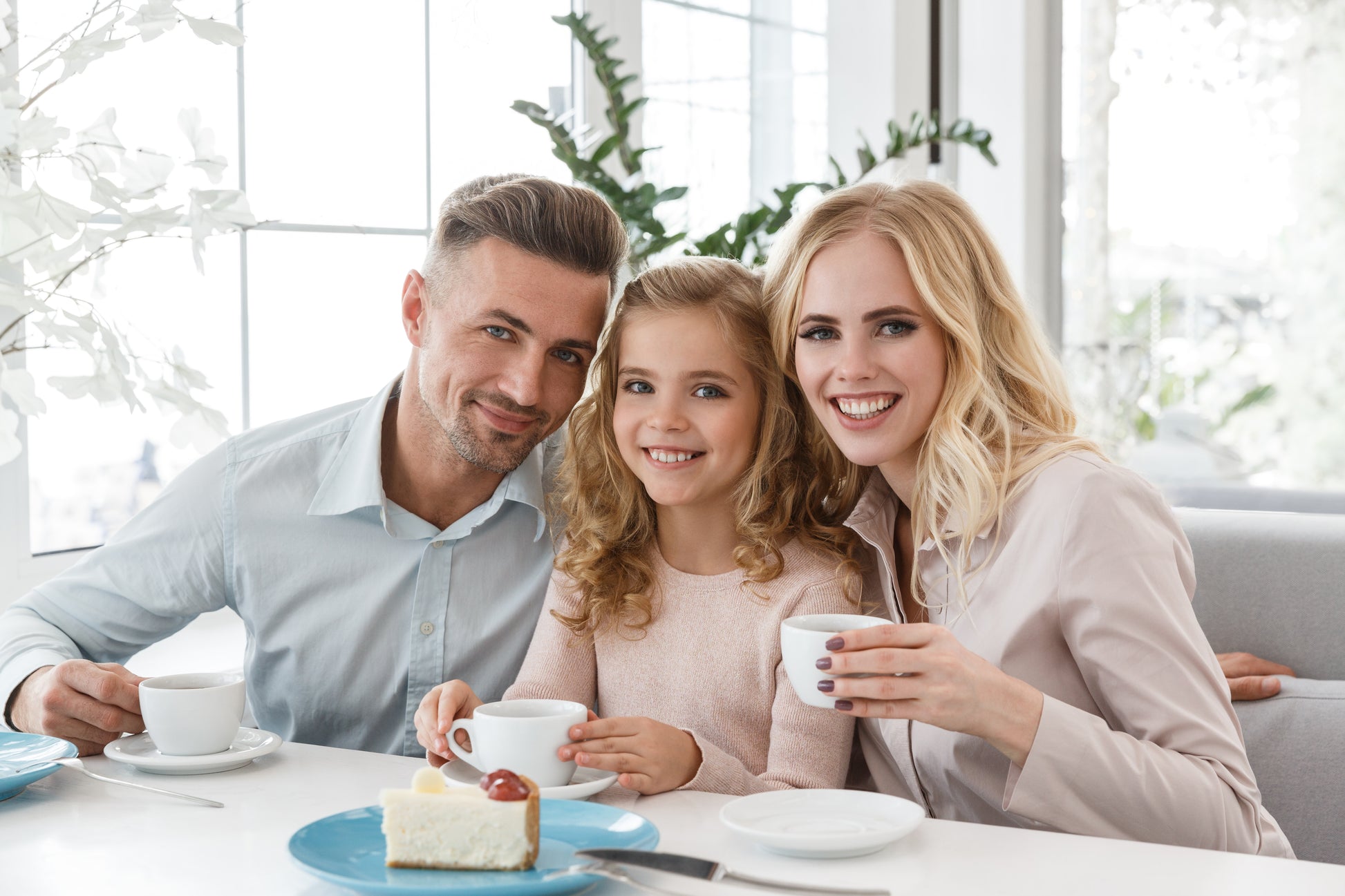 Family of three with parents drinking Ma' Cline's Coffee and daughter drinking a cup of ChocoShroom hot choclate. Aslice oof cheesecake with strawberry topping is in the foreground and the family is sitting close together at their dining room table.