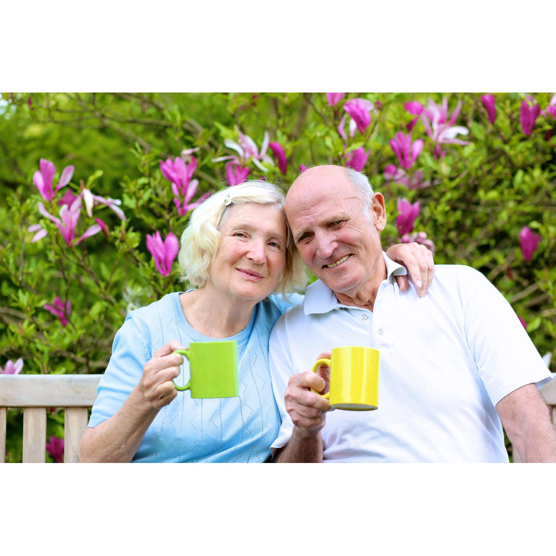 Elderly couple smiling, holding coffee mugs of delicious, healthy, Ma' Cline's Coffee, sitting on a bench in front of a blooming shrub with purple flowers.