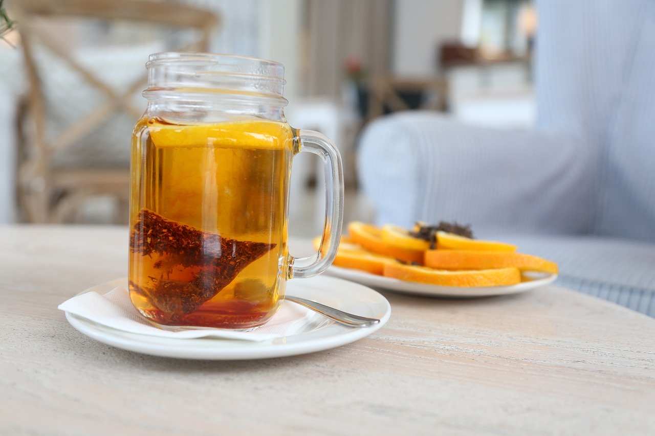 GanoCelebrate Rooibos tea in a transparent mason jar style cup on a white saucer with a napkin and a silver spoon. Behind is a plate of orange slices, all on a wood-grained table with a comfy overstuffed white chair in the background.
